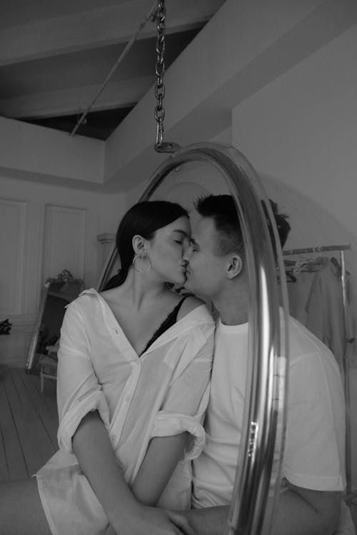 Black and white tender young couple wearing white shirts kissing and caressing each other while sitting on hanging egg chair in contemporary apartment