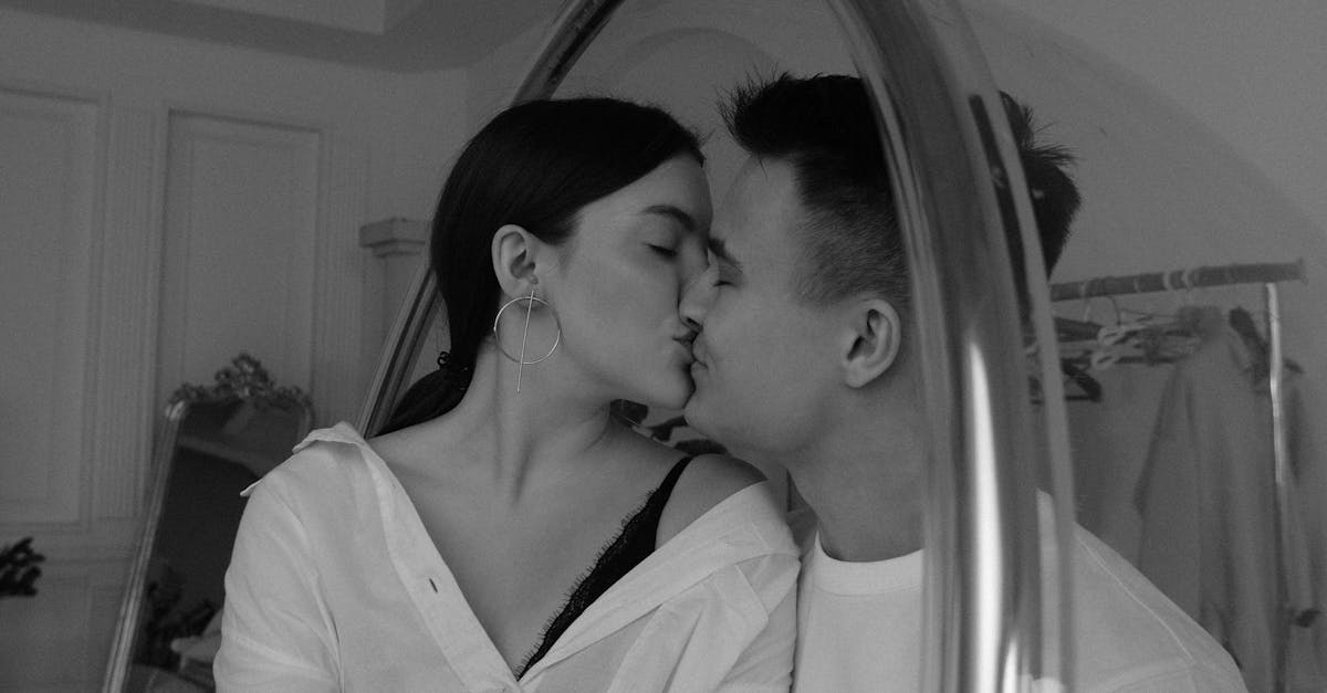 Black and white tender young couple wearing white shirts kissing and caressing each other while sitting on hanging egg chair in contemporary apartment