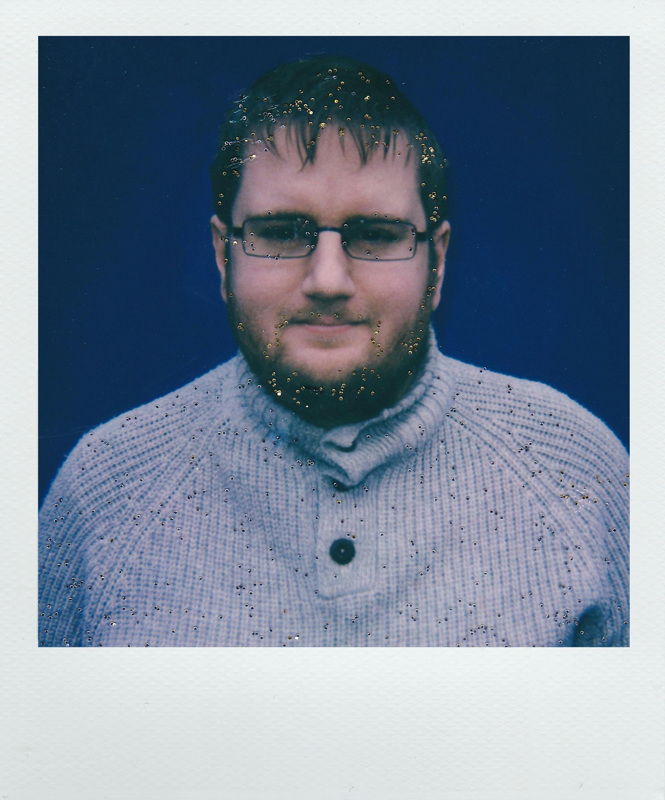 A bearded man with eyeglasses and a sweater smiles at the camera, accented by a glitter effect.