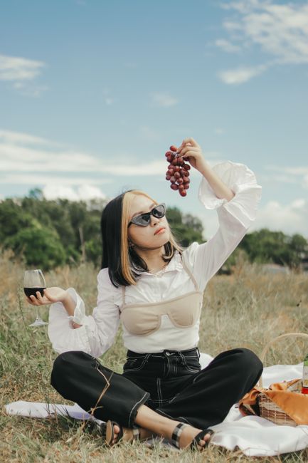 A woman, dressed in stylish attire, enjoys a picnic outdoors with wine and grapes.