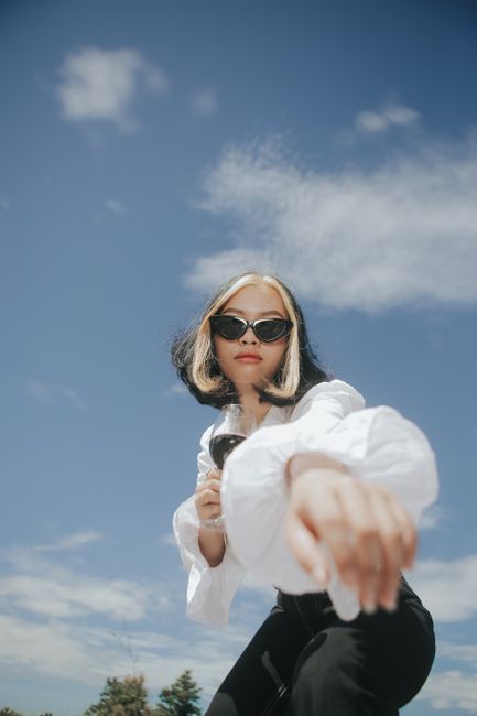 Fashionable woman in sunglasses and white blouse poses with a wine glass against a blue sky.