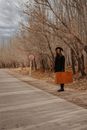 Full body of female in elegant outfit and hat with old suitcase near leafless trees in autumn
