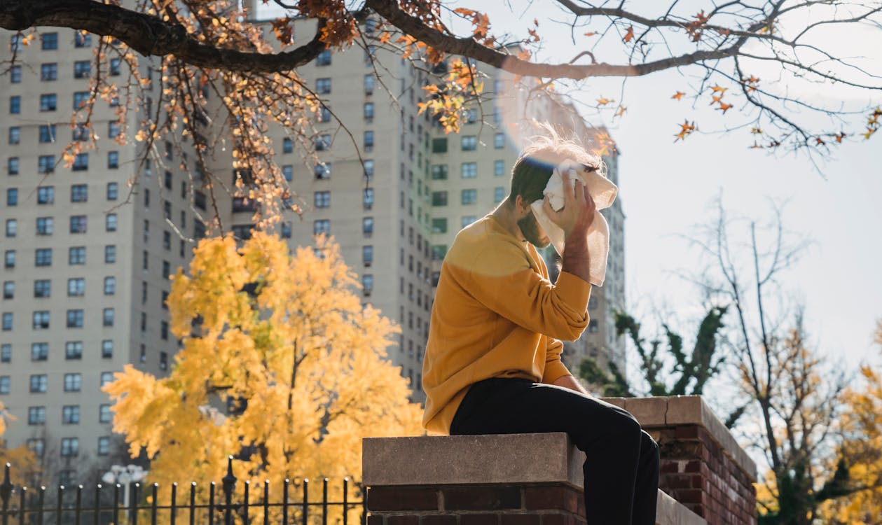 Side view unrecognizable male in orange hoodie sitting on brick border in city park and wiping sweat from forehead with towel