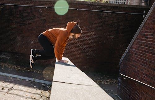 Side view full body energetic young male tracer in hoodie and hat jumping over barrier against brick wall on sunny autumn day