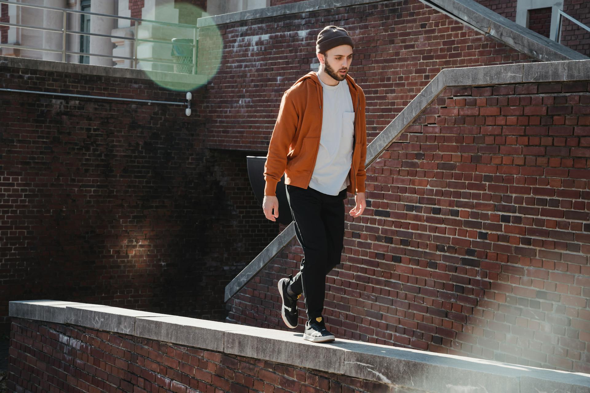 Full body young concentrated male in casual wear walking on narrow parapet of brick building on sunny day