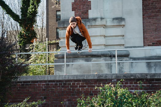 Full body determined young male tracer wearing hoodie and hat jumping over metal railing near old break building in sunlight