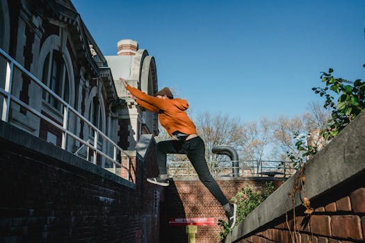 Side view full body fit male tracer wearing orange hoodie jumping from brick parapet towards building railing on clear day