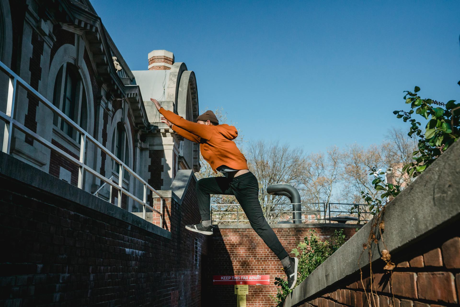 Side view full body fit male tracer wearing orange hoodie jumping from brick parapet towards building railing on clear day