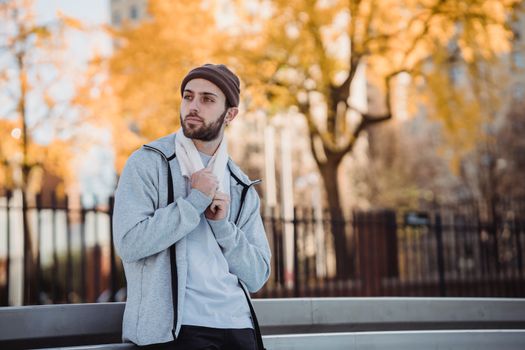 Thoughtful guy in trendy outwear standing on street against fence and tree with autumn leaves and looking away in daytime