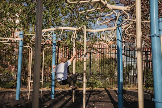 Back view of adult man in casual outfit hanging on metal pipes during parkour workout on sports ground