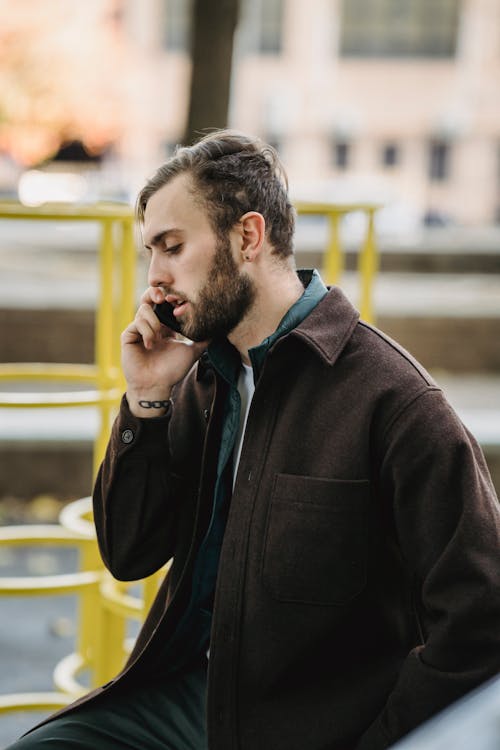 Side view of young bearded man in suede jacket talking on smartphone on playground