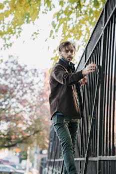 Young man wearing a stylish jacket, standing by a metal fence on a city street, showcasing urban fashion.