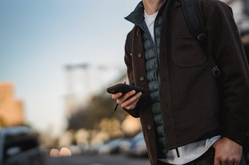 Crop anonymous male in casual outfit with cellphone on urban roadway under light sky