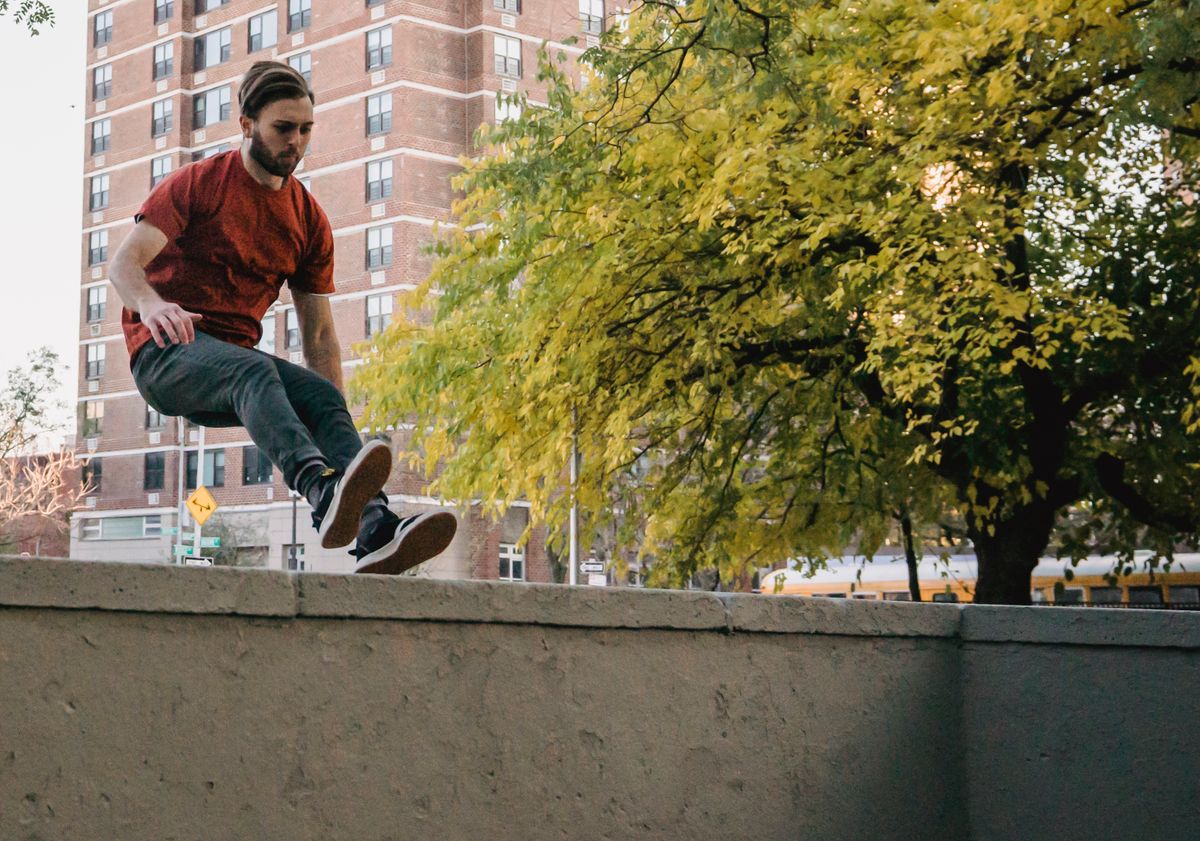 A traceur jumping over a concrete wall on a city street with trees and apartment buildings behind.