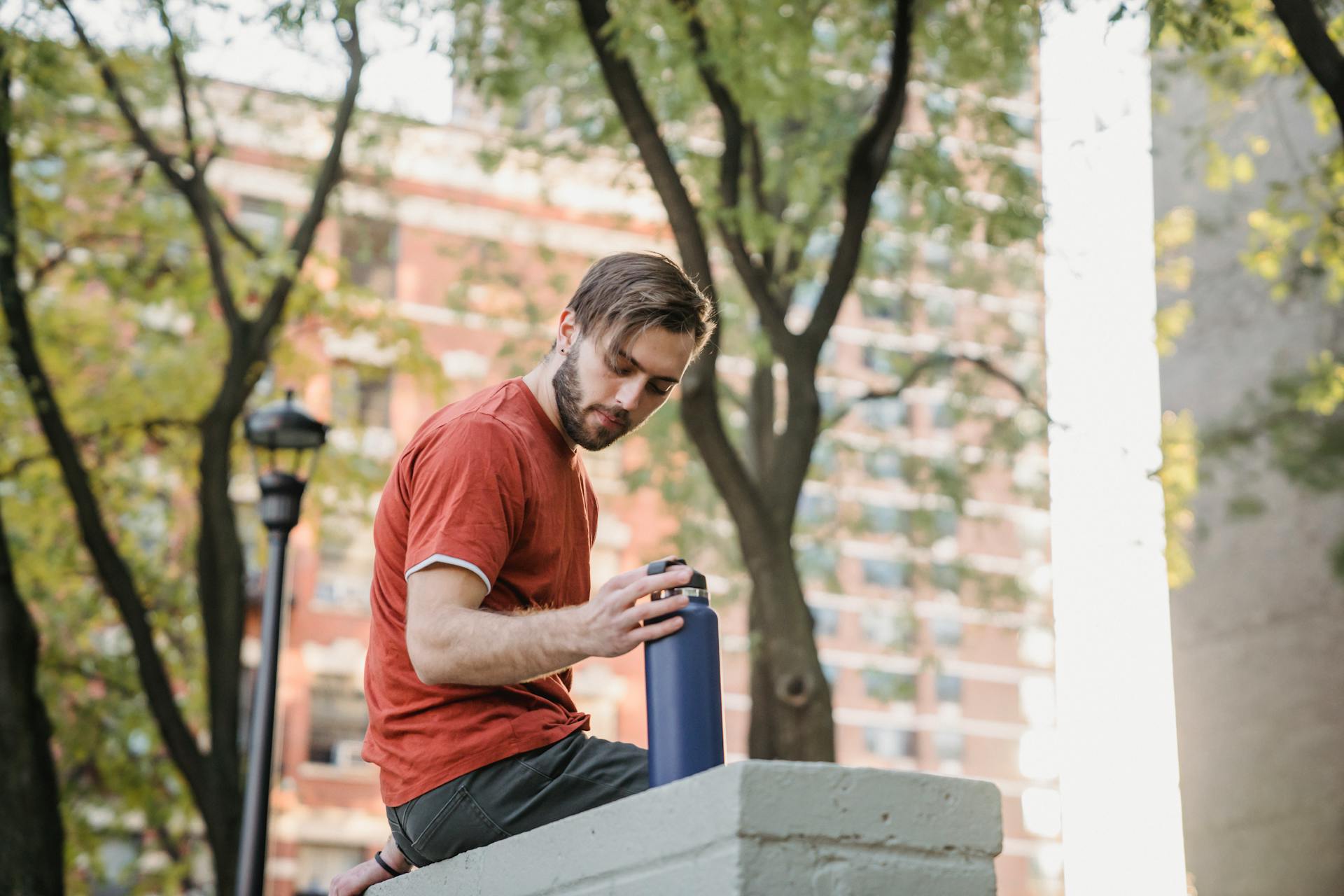 Low angle side view of young unshaven male athlete with bottle resting on fence in town