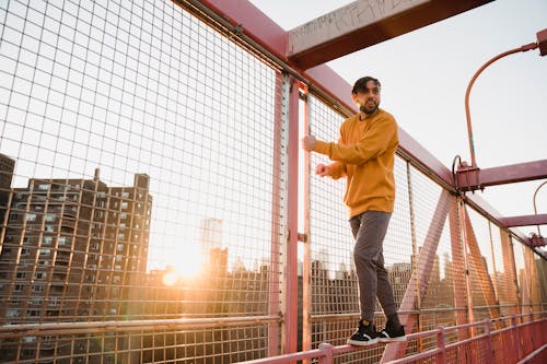 Free Full body of young bearded male in casual apparel standing on fence of urban bridge while looking away in sunshine Stock Photo