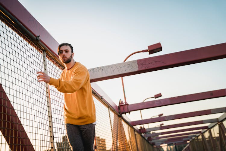 Attentive Bearded Man Near Grid Fence Of Bridge