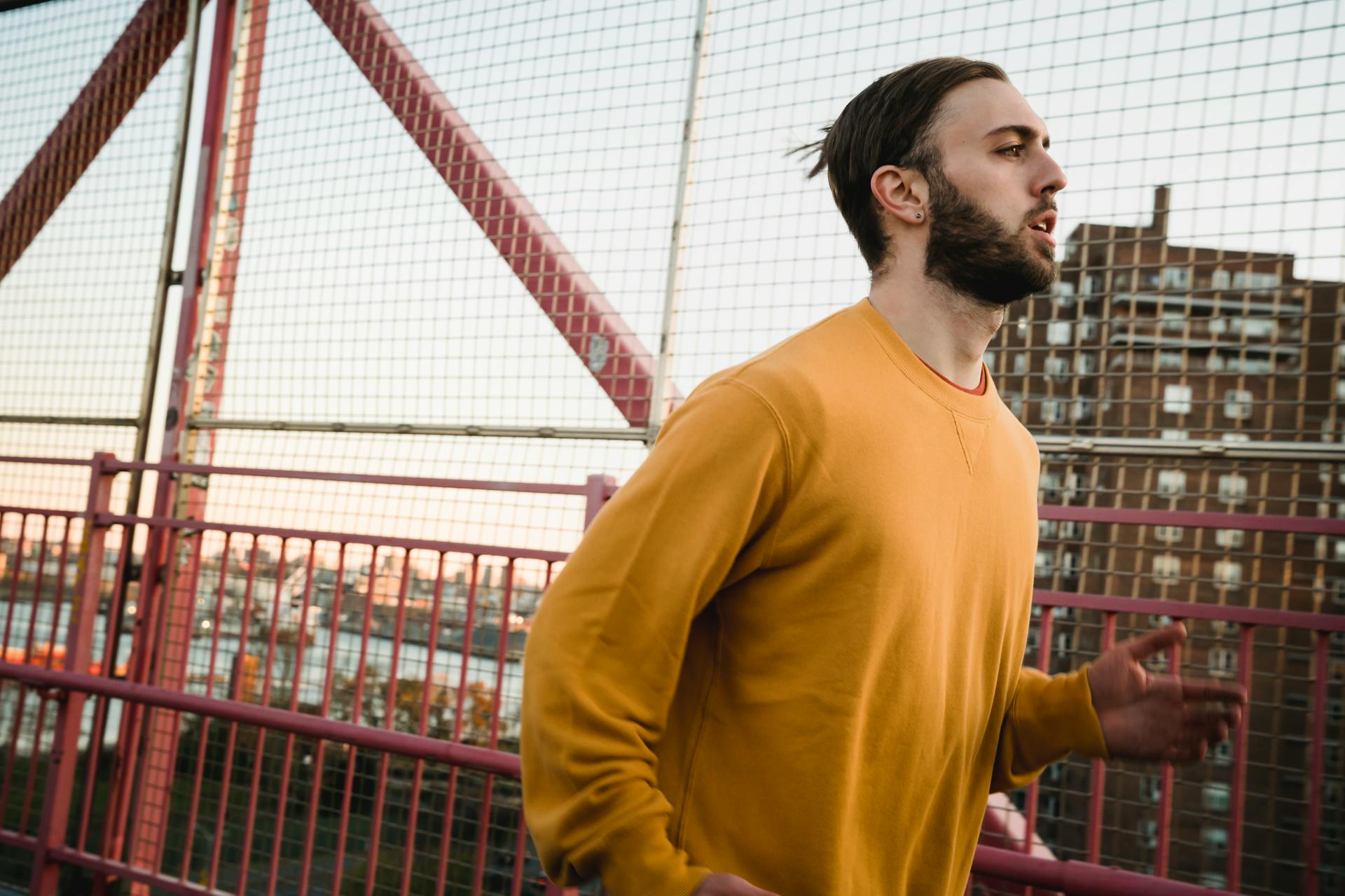 Side view of bearded sportsman running on pedestrian bridge along metal fence while training on street with building on background