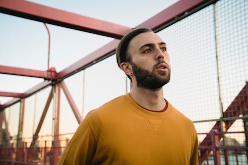 Sportive bearded male looking away while running on footbridge with metal red grid during training on street against blue sky