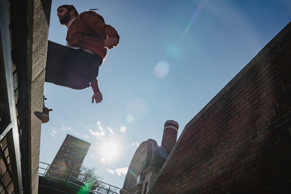Free From below of sportive fearless bearded male jumping on roof against blue sky in sunlight on street during parkour training Stock Photo