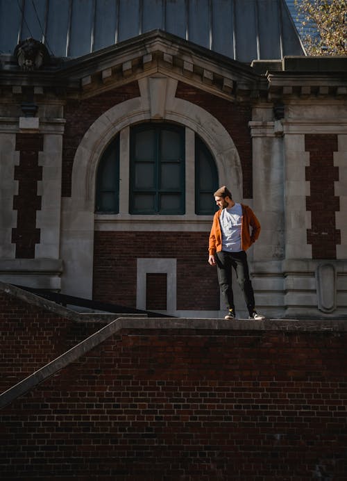 Free Full body of male wearing hat standing on brick wall with hand on waist on sunny street against historic building Stock Photo