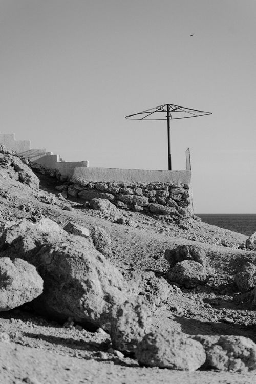 Free Black and white coastal scene with an empty umbrella frame atop rocky terrain. Stock Photo