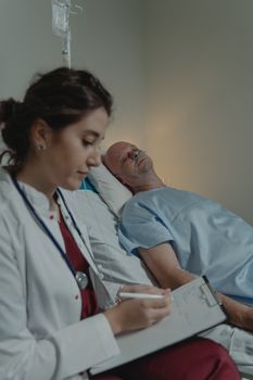 A doctor writes notes while attending to a hospital patient lying in bed, receiving care.