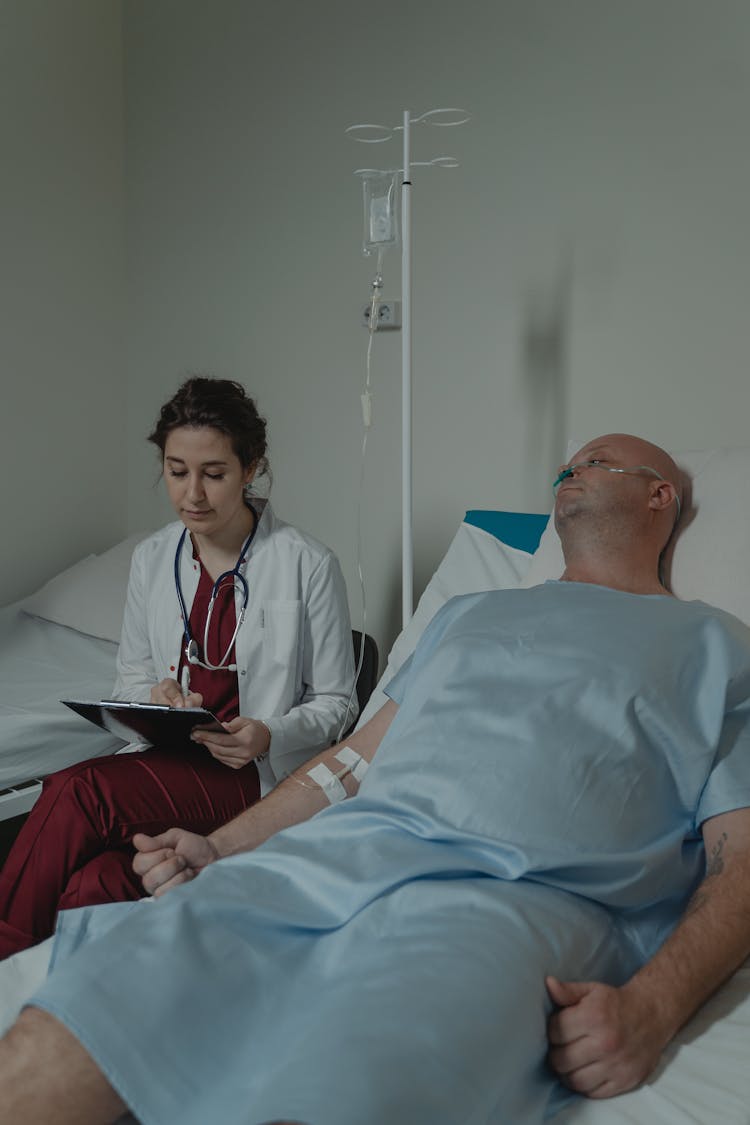 Doctor Writing On Medical Chart While Sitting Beside A Patient