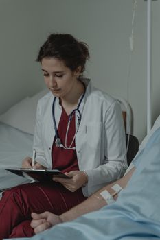 Medical professional with stethoscope taking notes beside patient in hospital setting.