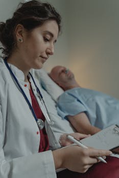 A female doctor takes notes beside a patient in a hospital room, showcasing medical care.