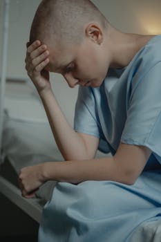 Bald woman in a hospital gown, sitting and looking down, symbolizes illness and contemplation.