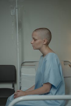 Bald woman in hospital gown sits pensively on a hospital bed.