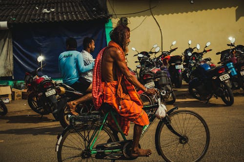 Side view of barefooted mature Indian male with long hair and beard in traditional clothes riding bicycle on asphalt road near unrecognizable men sitting on motorcycle at night
