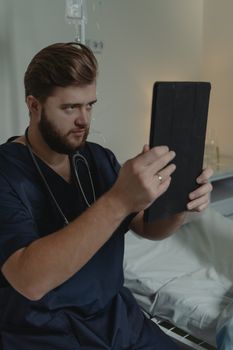 Healthcare worker with stethoscope using tablet, sitting by hospital bed, focuses on screen.