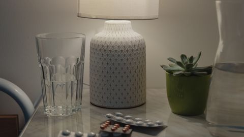 A warm nightstand setup featuring a lamp, glass of water, and medical tablets in a cozy bedroom environment.