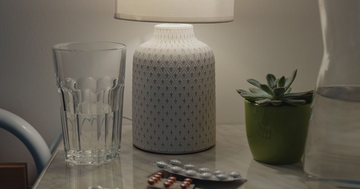 A cozy bedside nightstand at night with a lamp, a glass of water, a small succulent plant, and a blister pack of pills