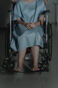 A young adult wearing a hospital gown sitting barefoot in a wheelchair indoors.