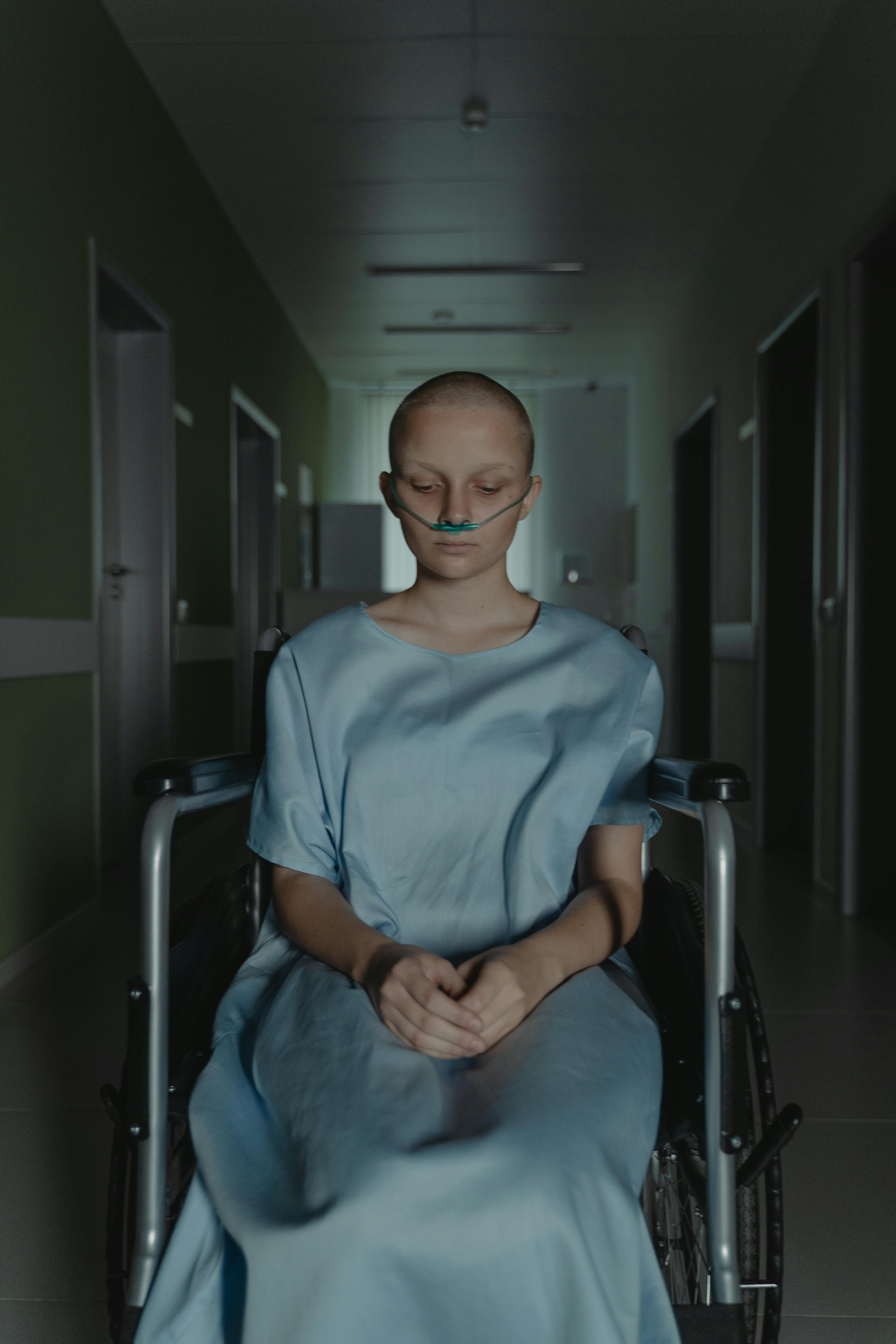 Woman in hospital gown sitting in wheelchair, conveying emotion in a dim hospital corridor.