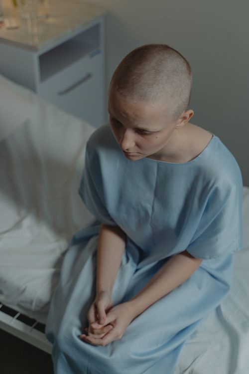 Free Bald woman in a hospital gown sitting on bed, expressing sadness and vulnerability. Stock Photo