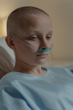 A bald woman wearing a nasal cannula in a hospital setting, reflecting a somber mood.