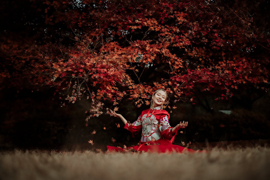 Cheerful ethnic female in traditional clothes smiling and catching red maples leaves near tree in field