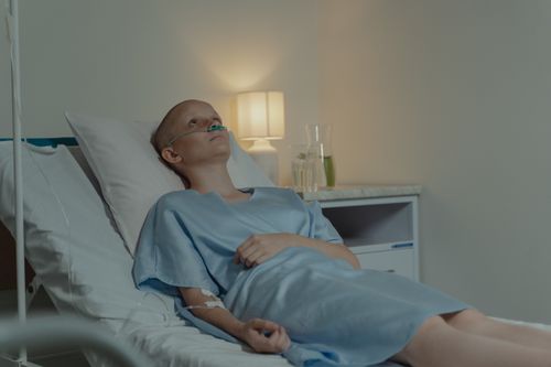 Free A woman in a hospital gown lying in bed with nasal oxygen support, reflecting on her condition. Stock Photo
