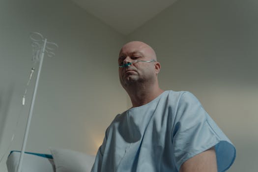 A bald man in a hospital gown sits in a room with a nasal cannula, conveying a sense of illness and solitude.