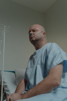 Bald man sitting in hospital room wearing a gown and nasal cannula, depicting illness.