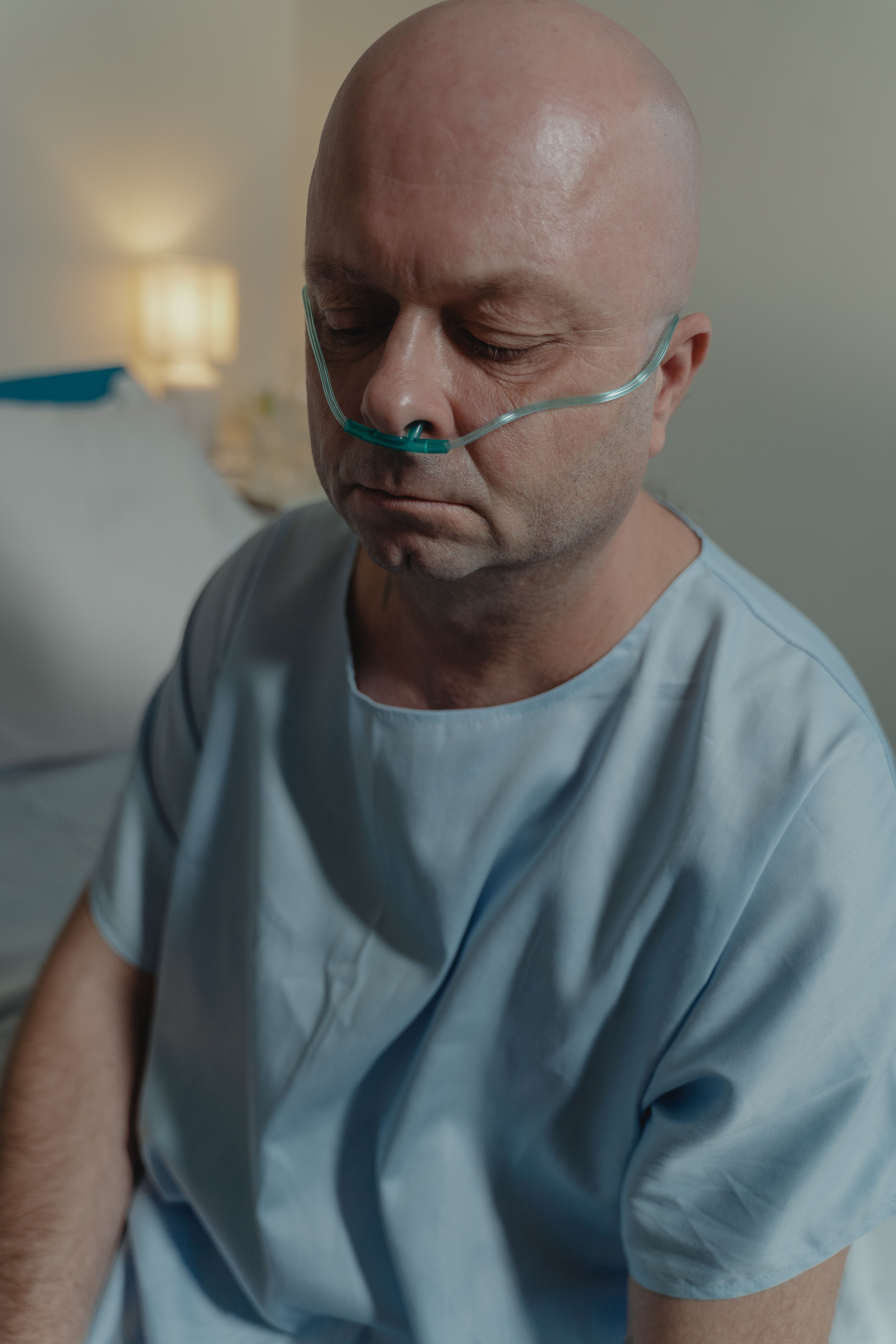 Bald adult man in hospital gown with nasal cannula, sitting indoors, conveying sadness and illness.