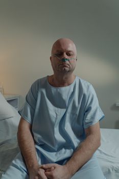 A bald man in a hospital gown sits on a bed, appearing thoughtful in a medical setting.