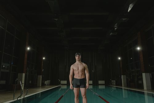 Free Fit male swimmer in swimming trunks and cap, standing poolside indoors. Perfect for sports and fitness visuals. Stock Photo