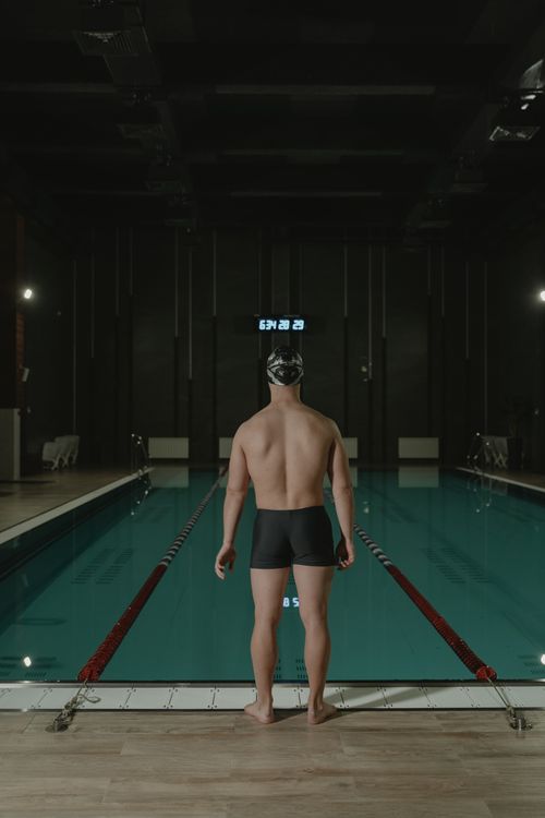 Free Male swimmer stands ready by the indoor swimming pool, showcasing athletic posture. Stock Photo