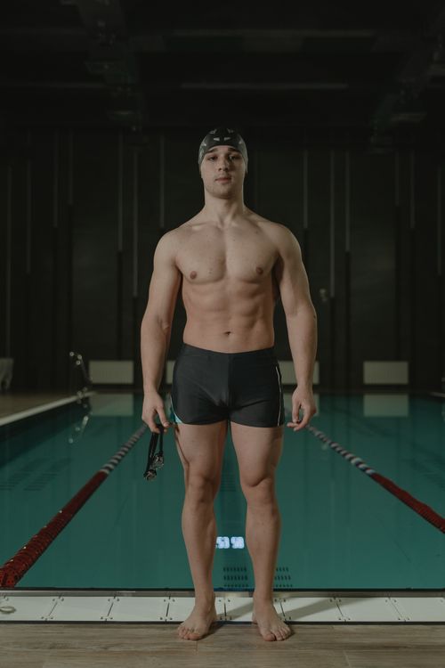 Free Adult male swimmer standing confidently poolside in swimming cap and trunks, showcasing athletic physique. Stock Photo