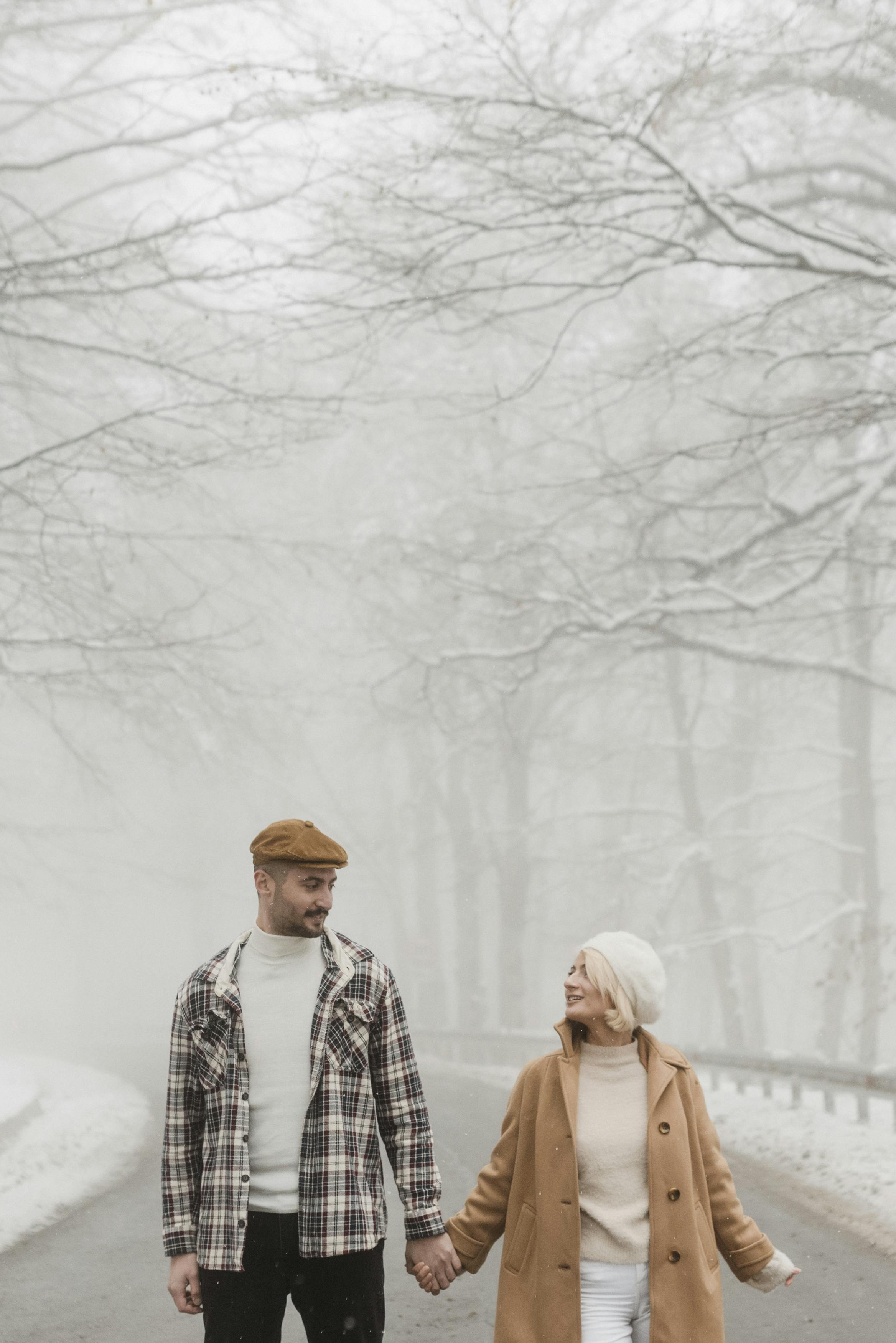 A couple holds hands while walking down a snowy road in a winter scene.